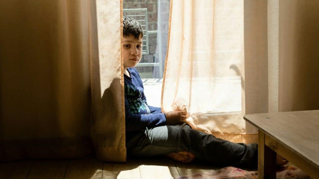 A young boy is sitting on a sunlit floor between two pulled-back beige curtains looking toward the viewer.
