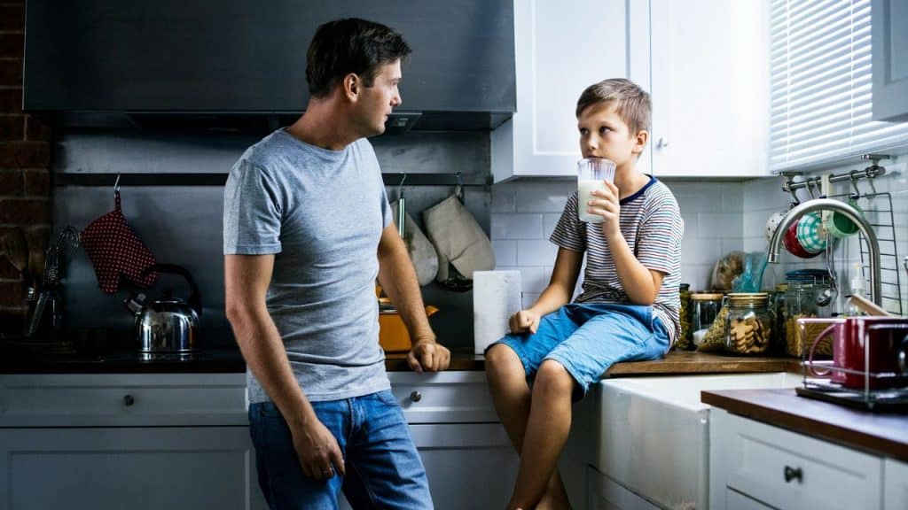 A man leaning on a kitchen counter speaks to a boy sitting on the counter, who is drinking a glass of milk.