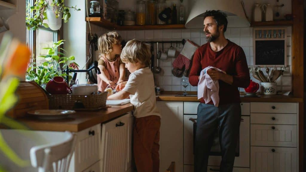 A man drying dishes stands in a sunlit kitchen while two young children interact at the kitchen sink.