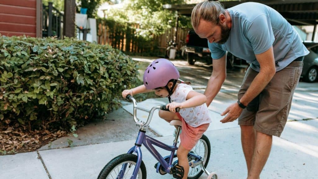 A man with a beard and ponytail helps a young girl wearing a purple helmet ride a small purple bicycle outdoors.