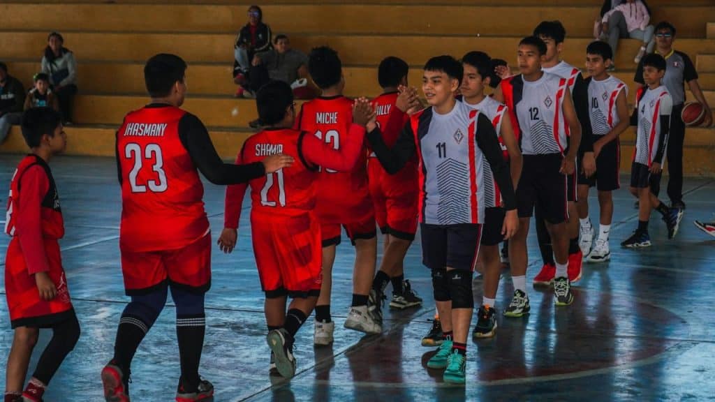 Young basketball players, some in red jerseys and others in white jerseys, line up and shake hands on an indoor court in front of a bleacher audience.