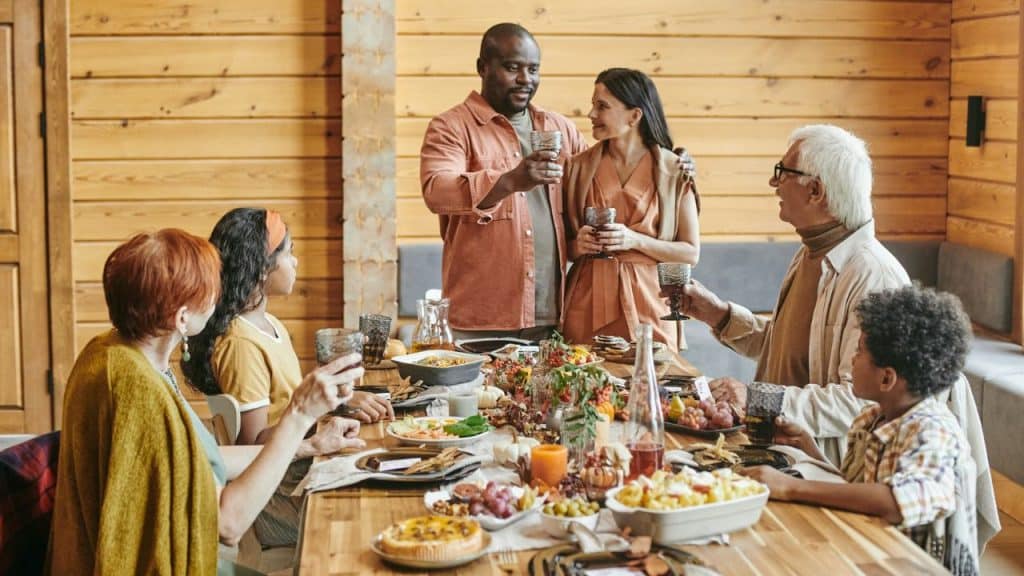 A multi-generational, multi-racial family gathered around a long wooden table with food is making a toast indoors.