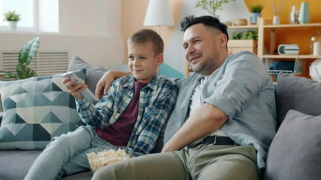 A smiling father and son are sitting on a couch with an arm around each other, watching television and eating popcorn.