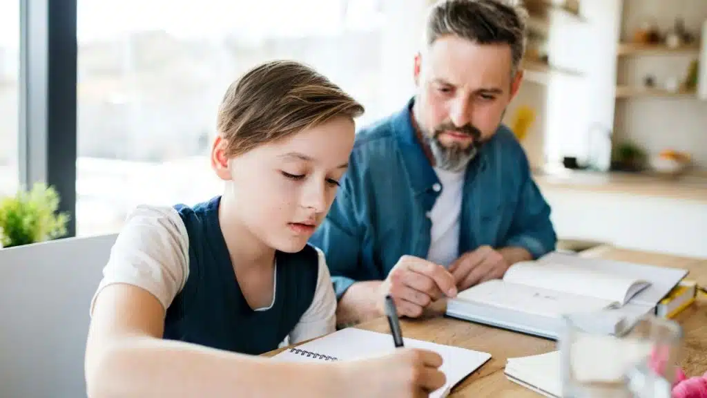 A father and son are sitting at a kitchen table with a boy writing in a notebook and the man watching him.