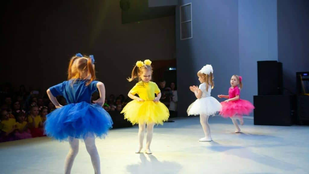 Four young girls in colorful tutu costumes are performing a dance on a stage in front of a dark audience.