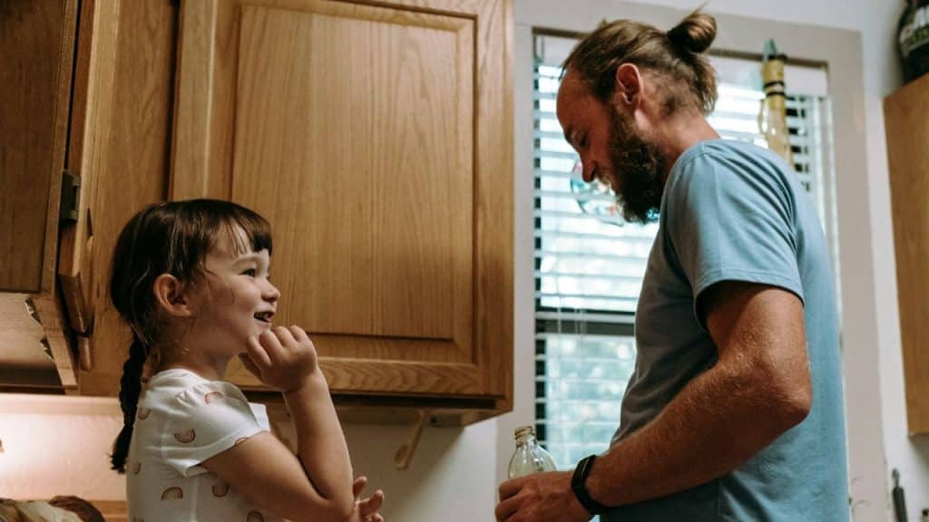 A bearded man with a bun and a small girl with a braid are smiling and talking in a kitchen.