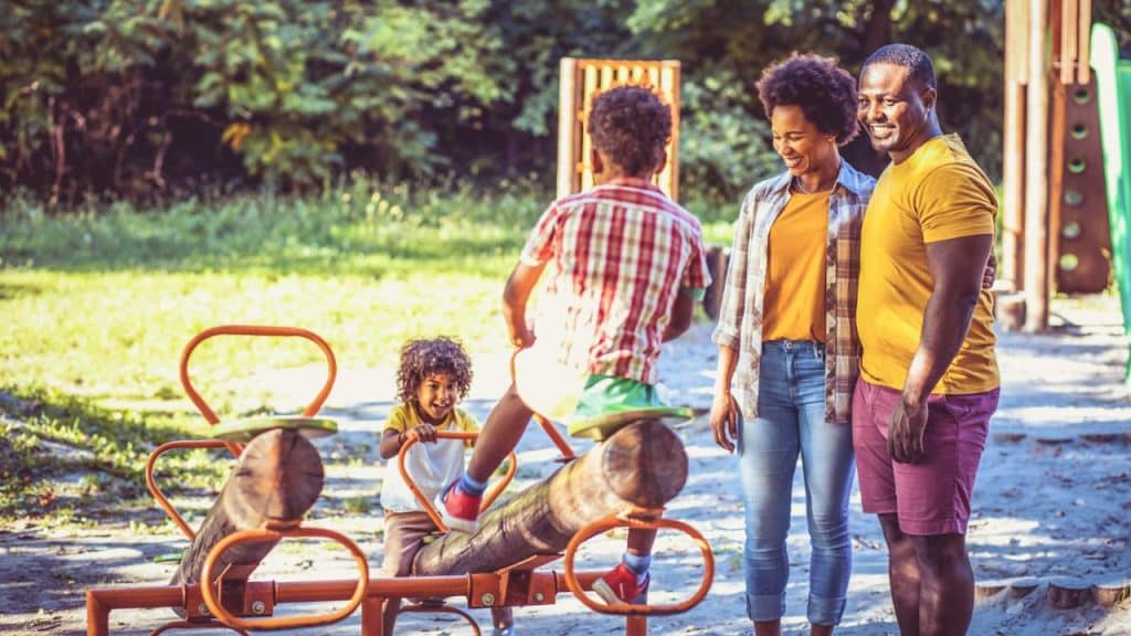 A Black couple smiles at a playground while two young Black children play on a wooden and orange metal seesaw.