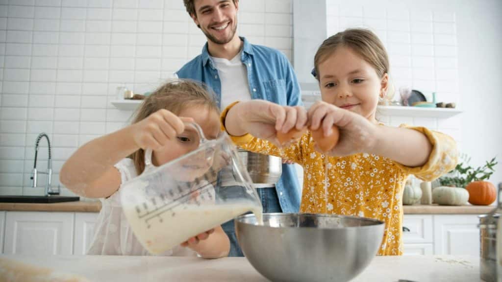 A man smiles in a kitchen while two young girls pour and crack ingredients into a metal mixing bowl.