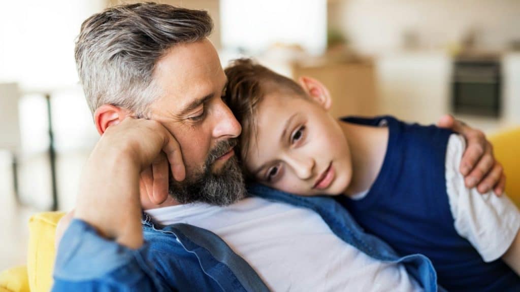 A bearded man is leaning back on a yellow couch while a young boy with short hair rests his head on his shoulder.