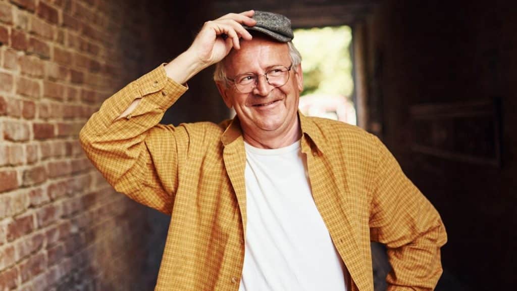 A smiling older man in glasses and a yellow plaid shirt tips his cap.