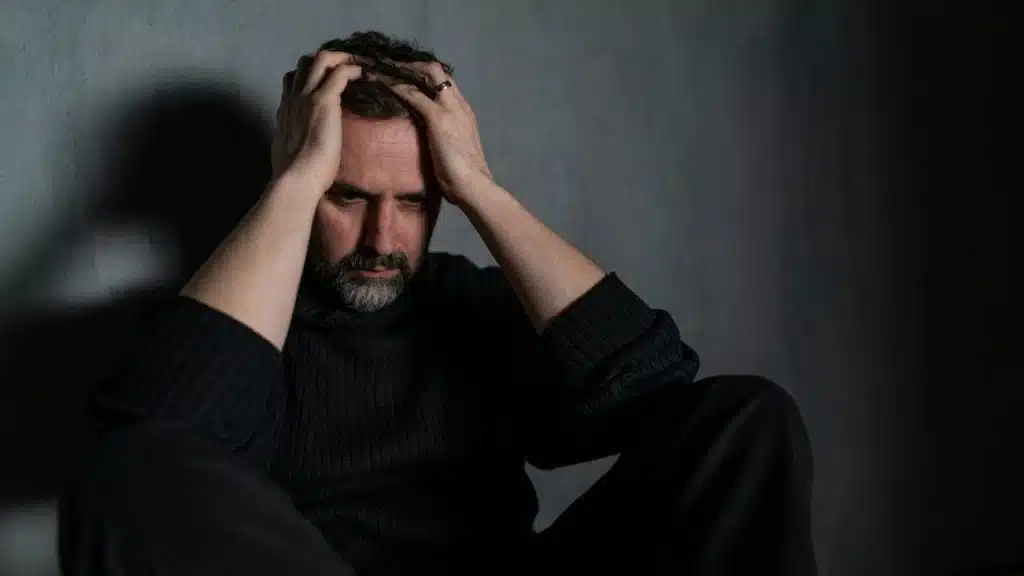 A stressed, bearded man in a dark turtleneck sits against a gray wall, holding his head in his hands.