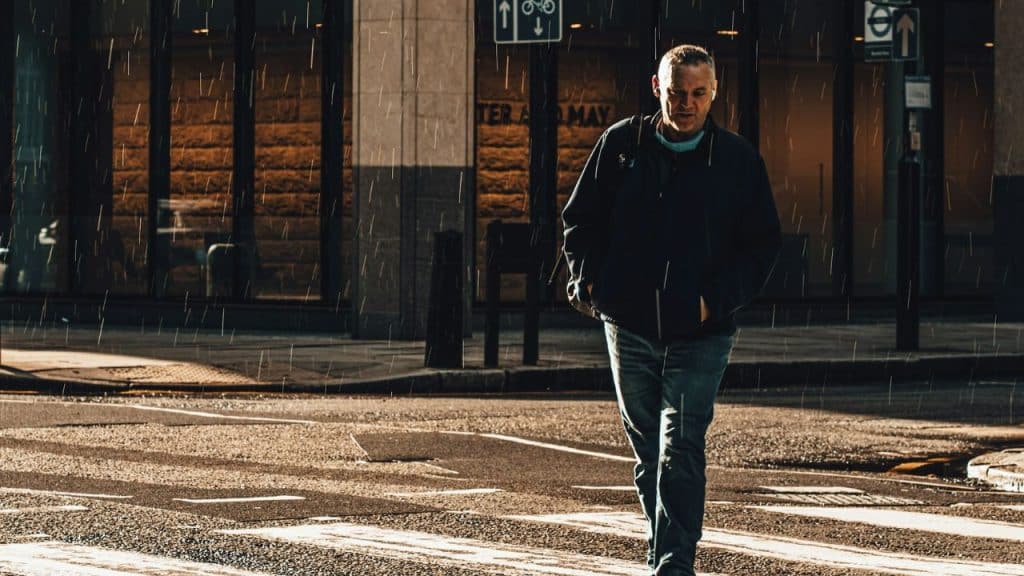 A somber, older man walks across a wet city crosswalk in the rain.