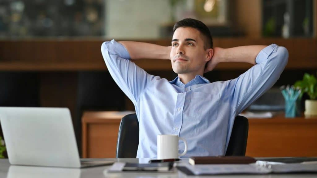 A content young man leans back in his office chair with his hands behind his head.