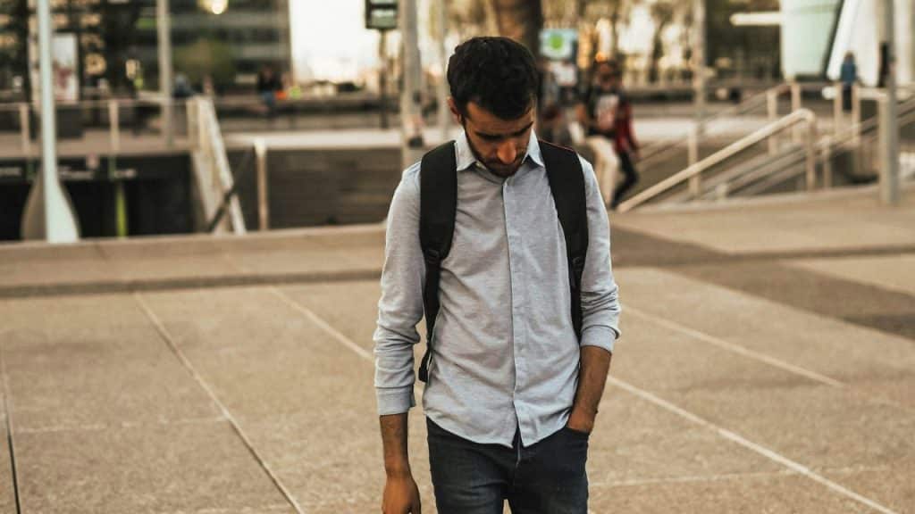 A disappointed young man with a backpack walks alone in a city plaza, looking down.