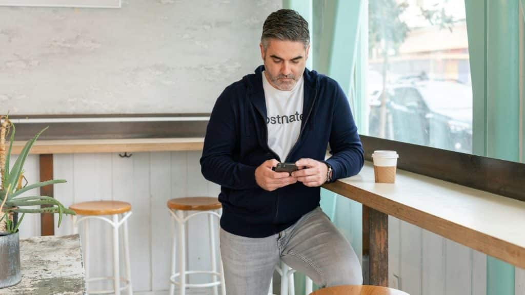 A casually dressed middle-aged man stands at a coffee shop counter, looking at his smartphone.