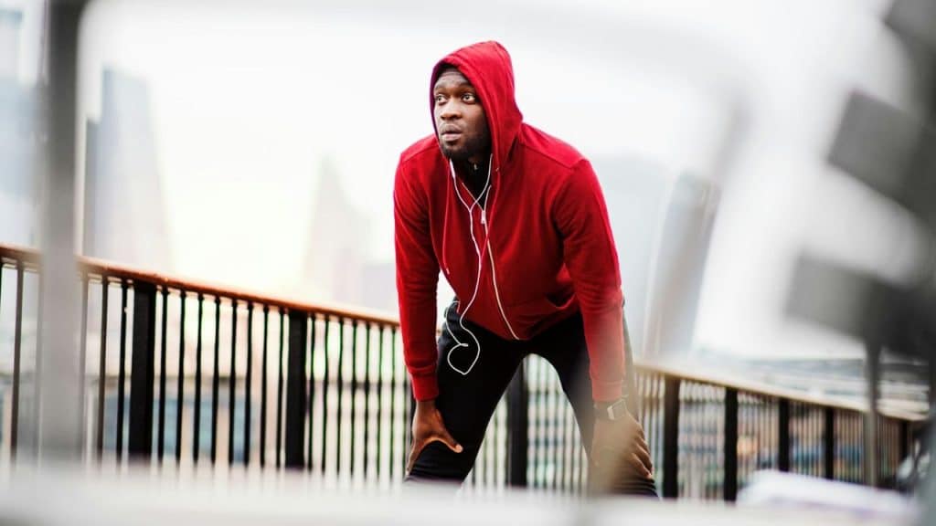 A Black man in a red hoodie and earbuds leans over, catching his breath after a run.