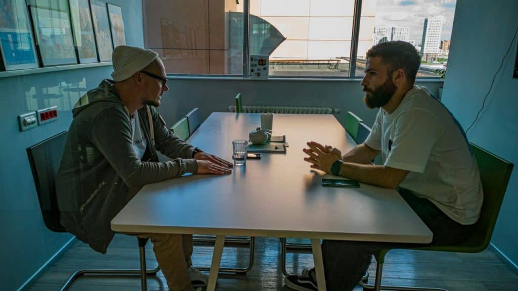 Two men sit at a long table, leaning in for a serious conversation in a modern office.