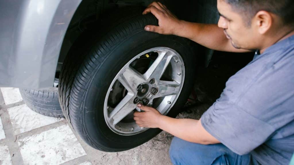 A male mechanic kneels, checking the lug nuts on a car tire in a garage.
