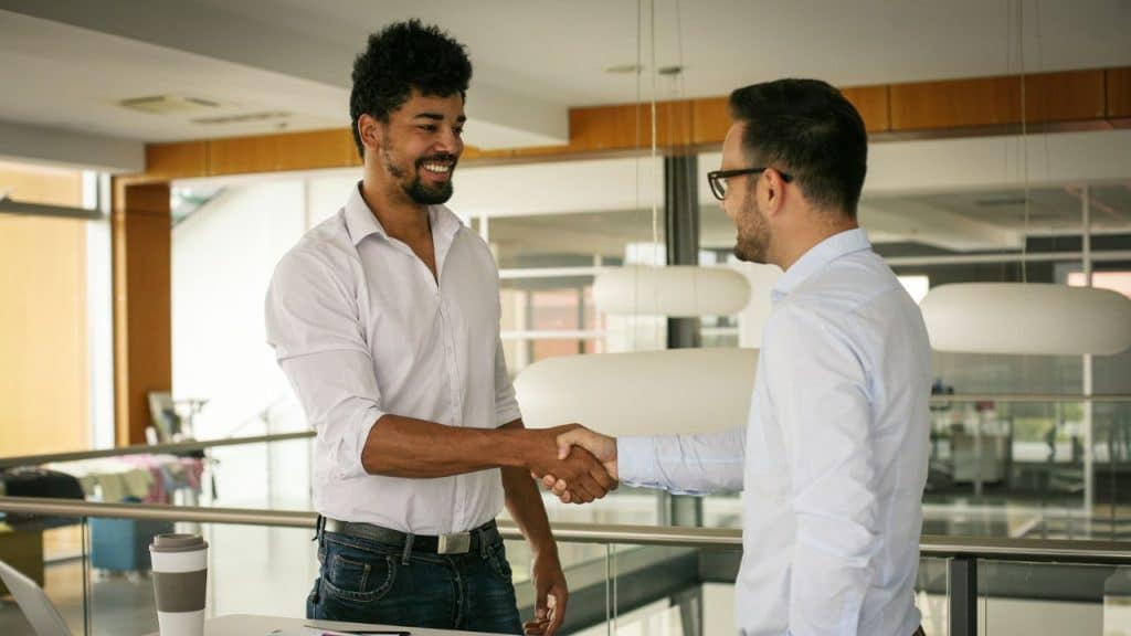 Two businessmen in dress shirts smile and shake hands in a modern, brightly lit office.