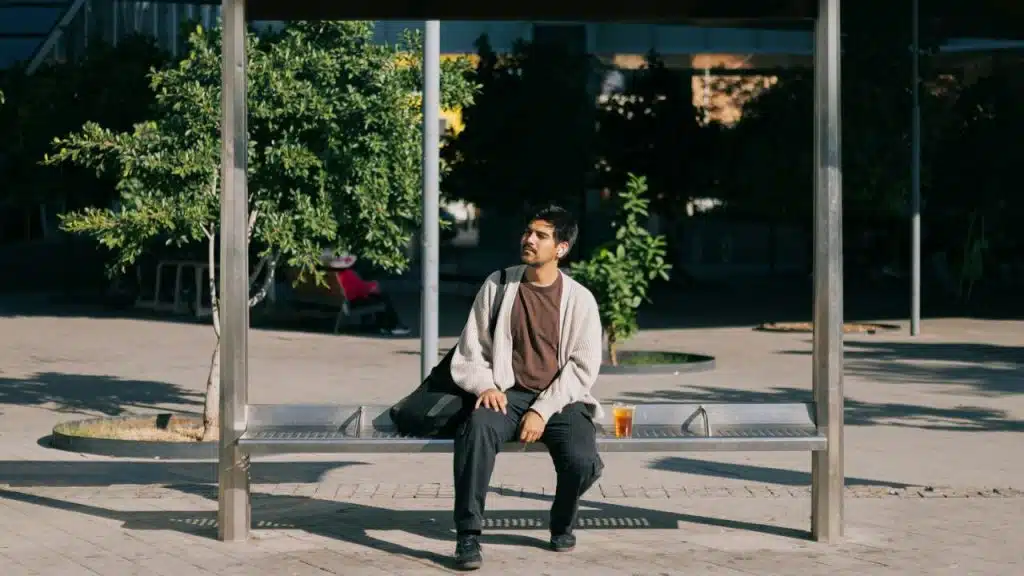 A young man in a brown shirt and cardigan sits alone at a sunny bus stop.