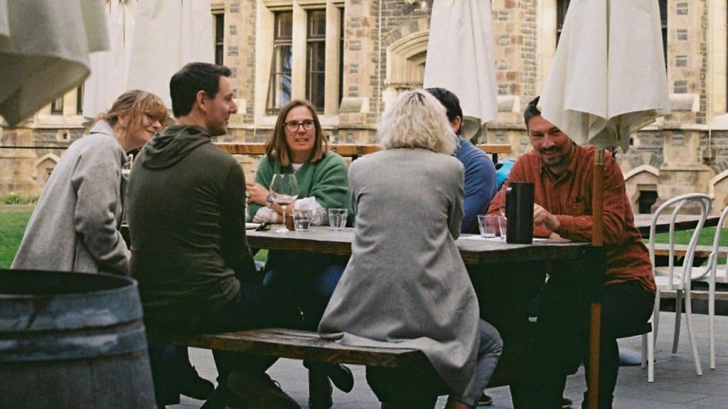Six people sitting around a rustic wooden table under umbrellas outside a stone building.