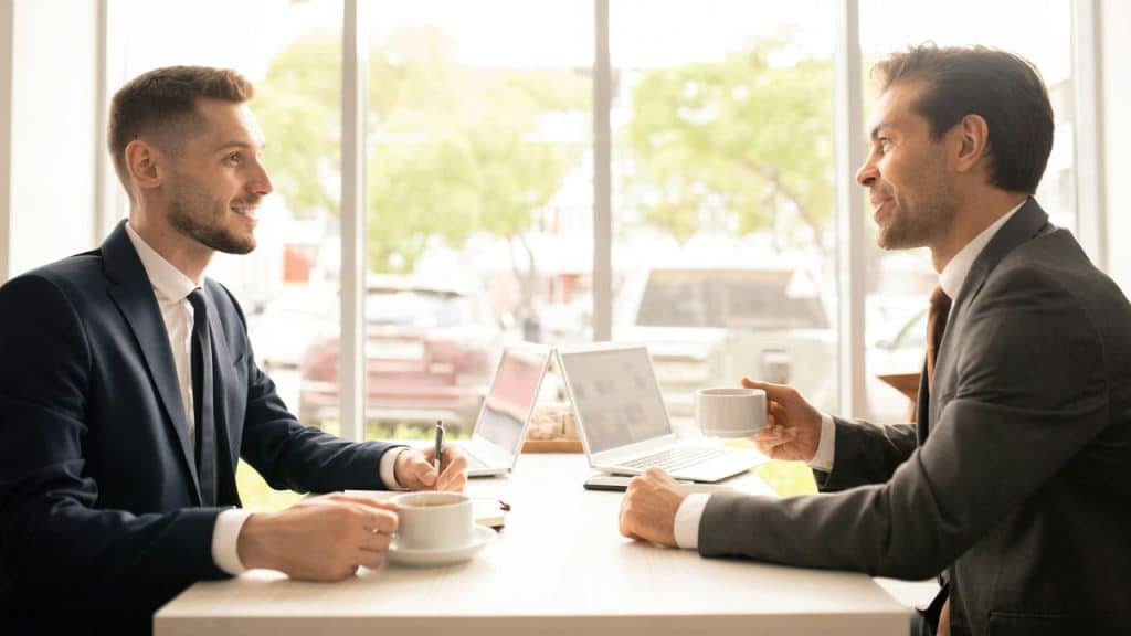 Two well-dressed businessmen sit across a table, smiling and talking while drinking coffee.
