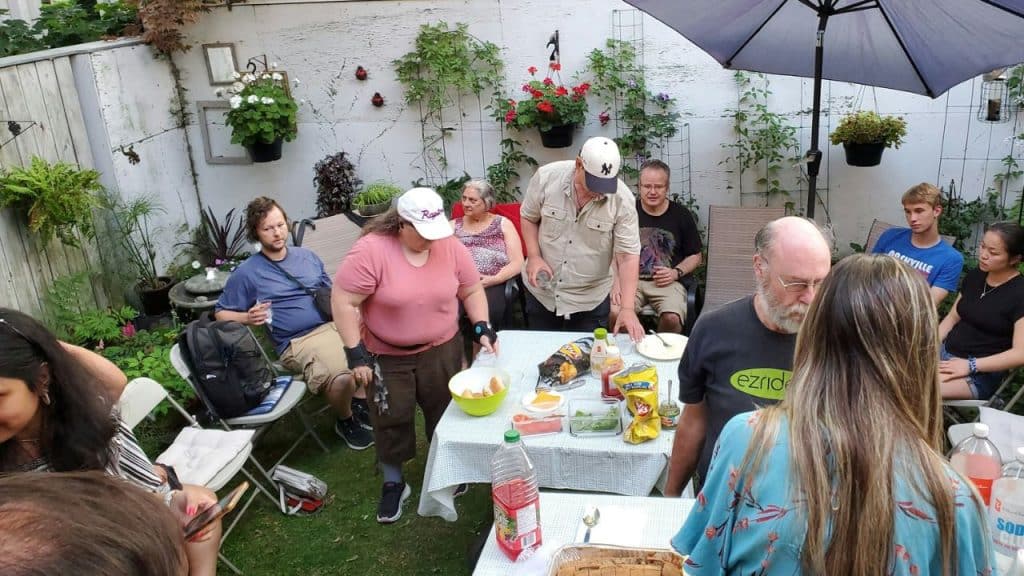 A large, multi-generational group of people are gathered around a food table in a backyard garden patio.