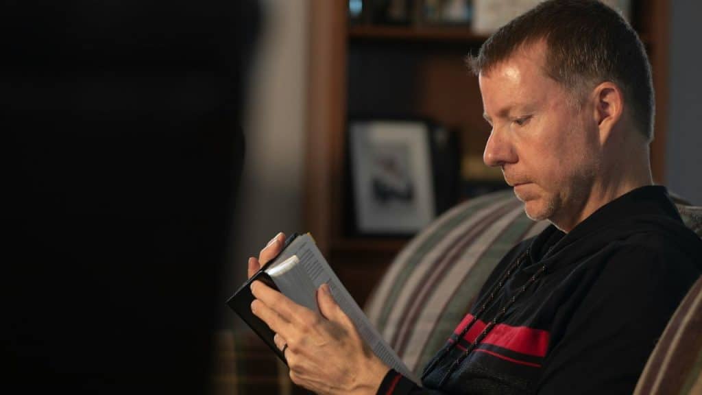 A focused man in a dark hoodie sits on a striped couch, reading a book.