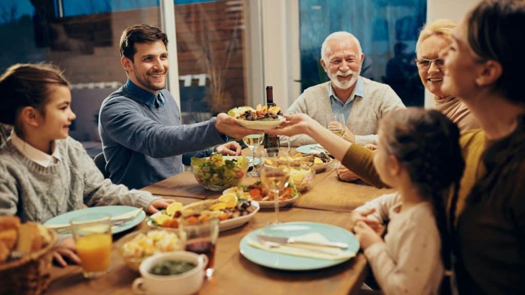 A happy, multigenerational family passes a plate of food during a dinner gathering.
