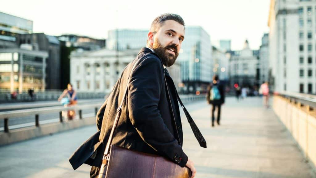 A well-dressed bearded man walks across a city bridge, looking back over his shoulder.