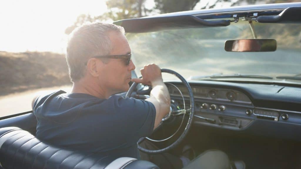 A gray-haired man in sunglasses drives an older convertible on a sunny day.