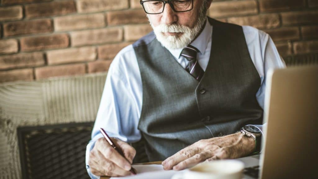 A serious, well-dressed older man with a white beard writes on paper with a pen.