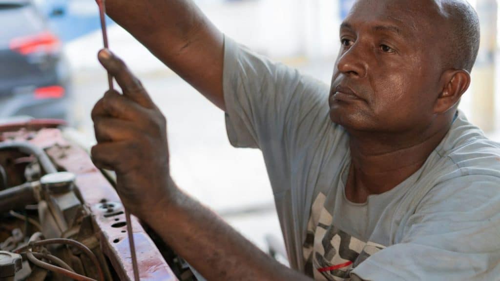 A bald man with greasy hands works on a car engine in a light blue t-shirt.