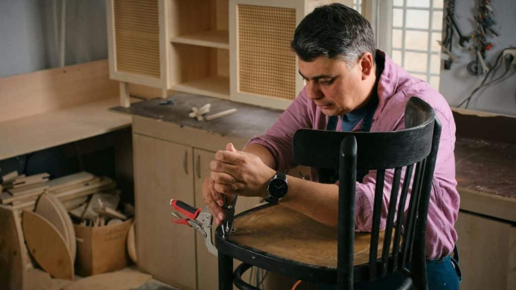 A craftsman in a pink shirt works with pliers to repair a wooden chair in a workshop.