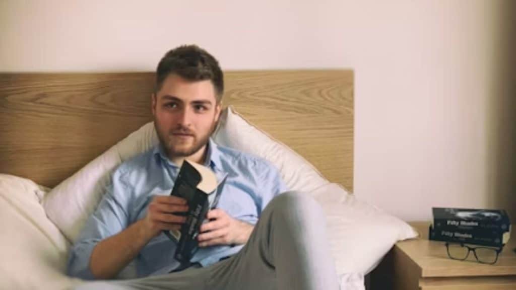 A man reading a book in bed with warm bedside lighting.