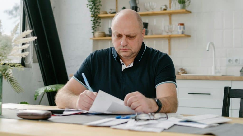 A balding man in a dark polo shirt sits at a table, writing on paper in a bright kitchen.