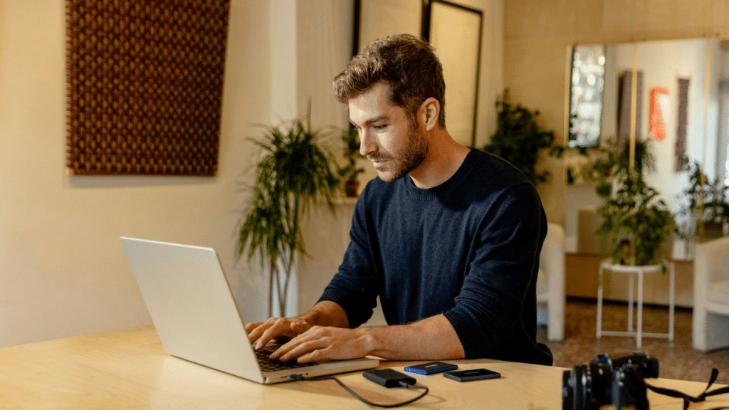 A serious, handsome man works on a laptop at a desk in a bright, modern room.