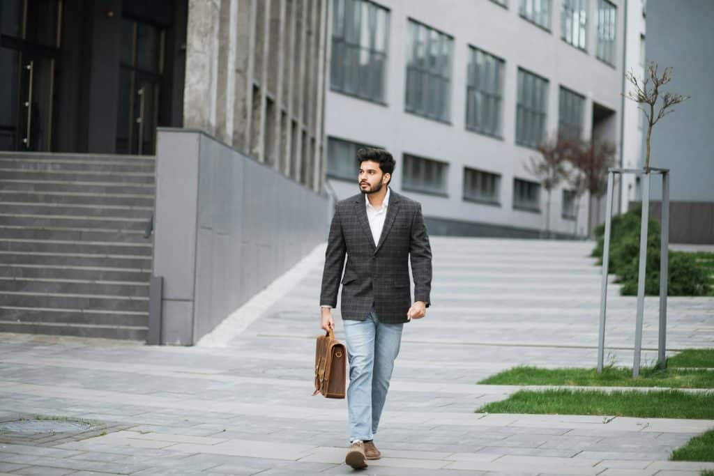 The man is walking outdoors carrying a brown leather suitcase.