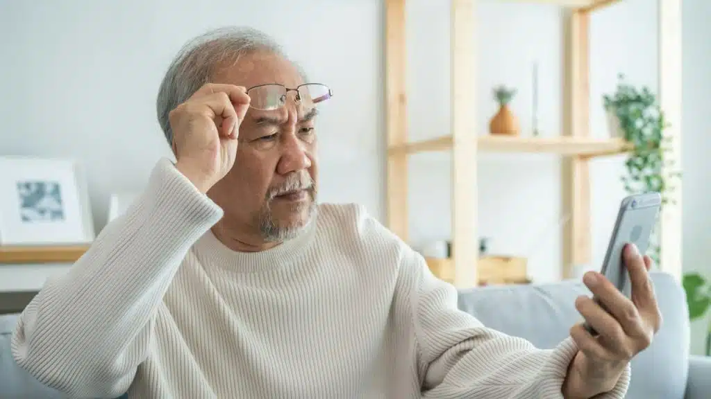 An older man squints, holding a smartphone and pushing his glasses up onto his forehead.