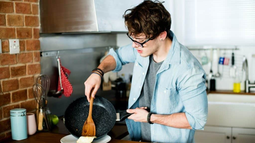 A young man wearing glasses and a light blue shirt cooks in a kitchen with a brick wall.