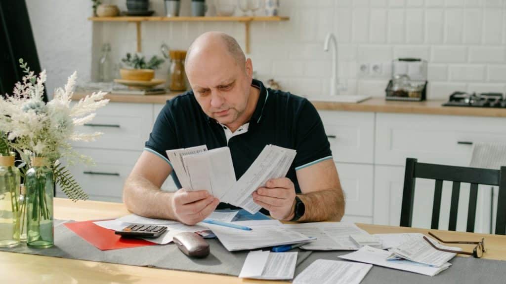 A balding man sits at a kitchen table, intently holding and reading a stack of paper bills.