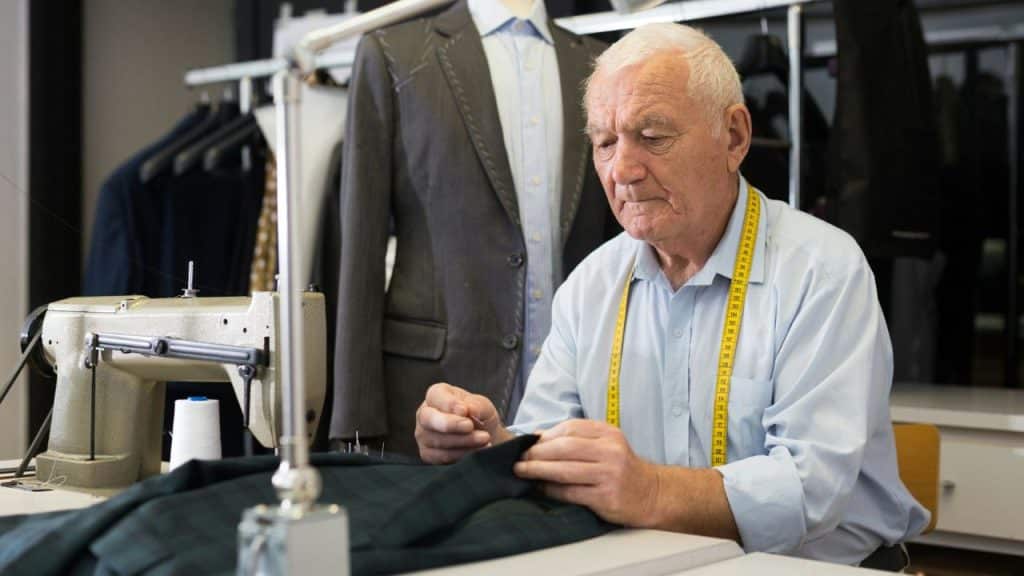 An elderly male tailor with a tape measure around his neck sews a dark piece of fabric.