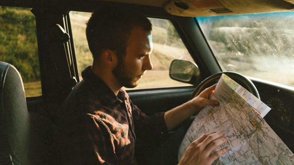 A young man with a beard sits in a car, seriously studying a folded paper map in sunlight.