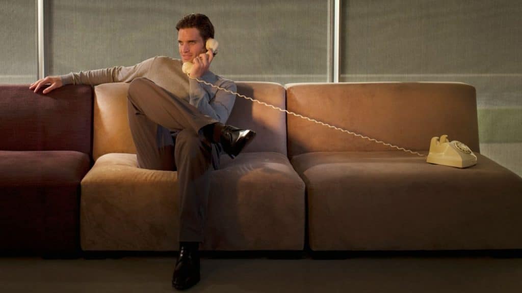 A well-dressed man sits on a sectional sofa talking on a yellow rotary phone with a long cord.