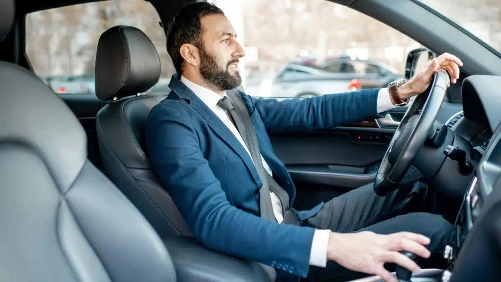 A smiling, well-dressed man with a beard sits in the driver's seat of a modern car.
