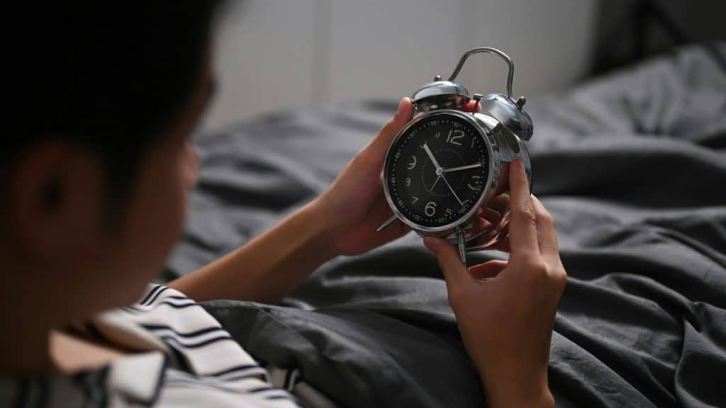 A person lying in bed holds and looks closely at a chrome twin-bell analog alarm clock.