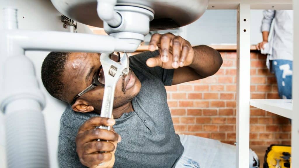 A man wearing glasses is underneath a sink, using a large adjustable wrench on a pipe.