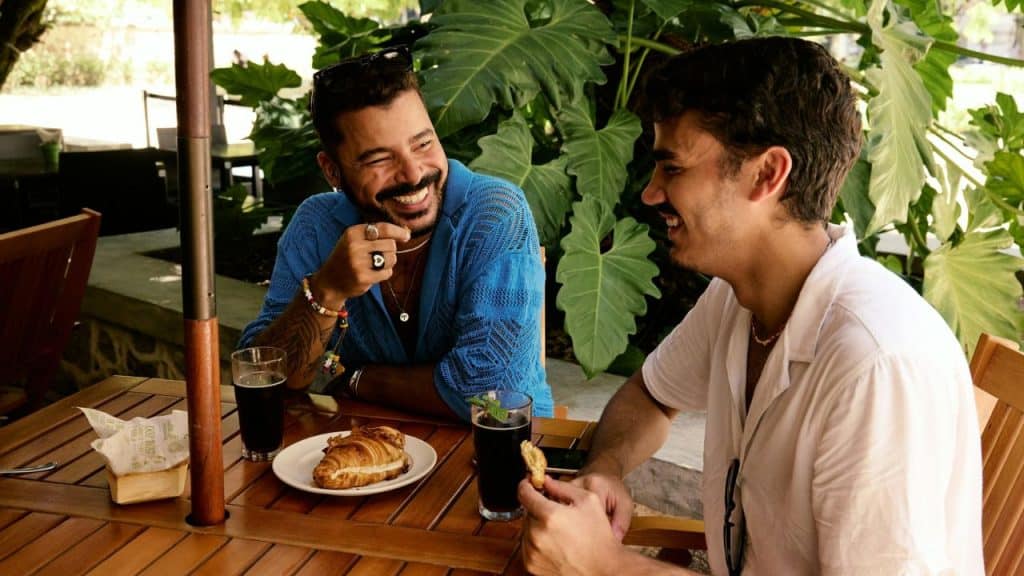 Two young men sit laughing together at a wooden outdoor cafe table with food and drinks.
