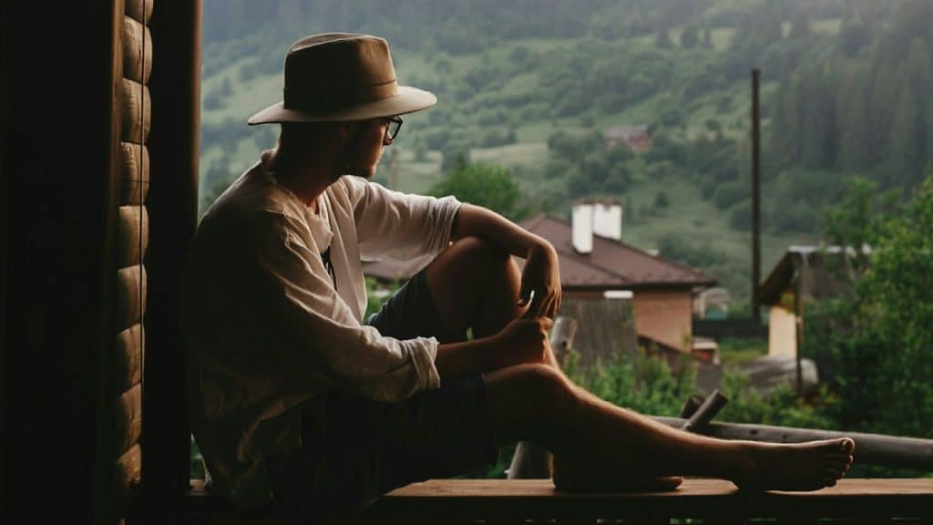 A man in a fedora sits barefoot on a porch, looking out at a hilly, green rural landscape.