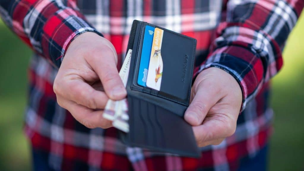 Hands in a red flannel shirt hold an open black wallet with cash and credit cards visible.